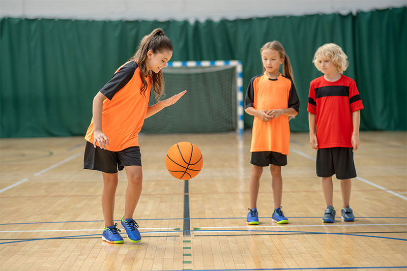 Girl dribbling a basketball on the basketball court wearing an orange jersey while two other players watch