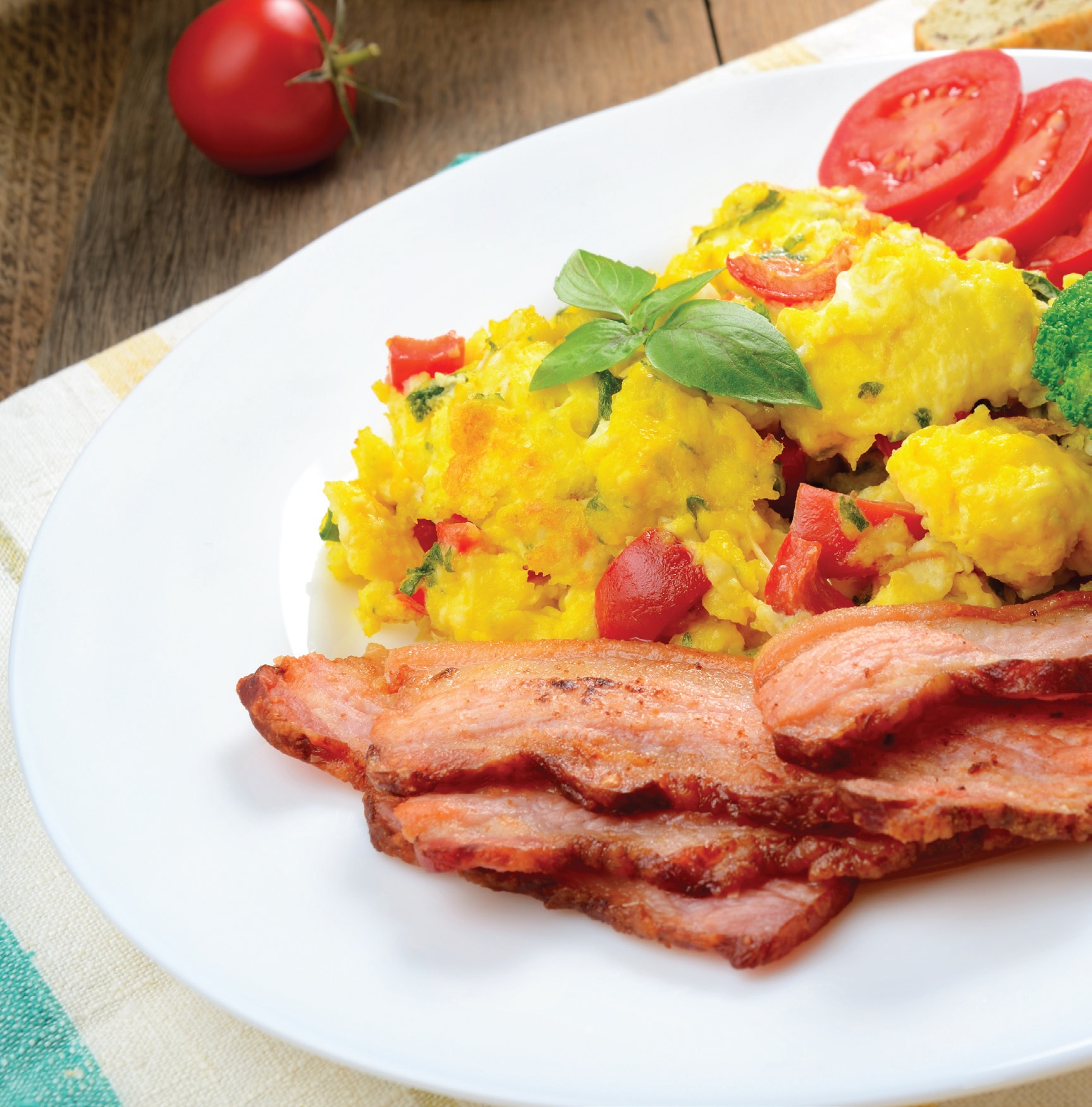 Scrambled eggs on a plate topped with tomatoes and basil next to slices of bacon