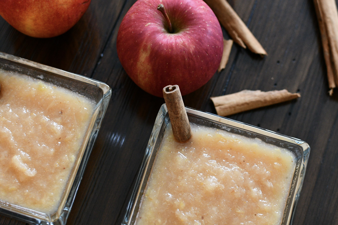 Homemade apple sauce in square glass bowls with cinnamon sticks and Washington red apples on a dark top