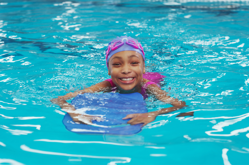 Girl swimming with googles on her head and holding a kickboard in the swimming pool