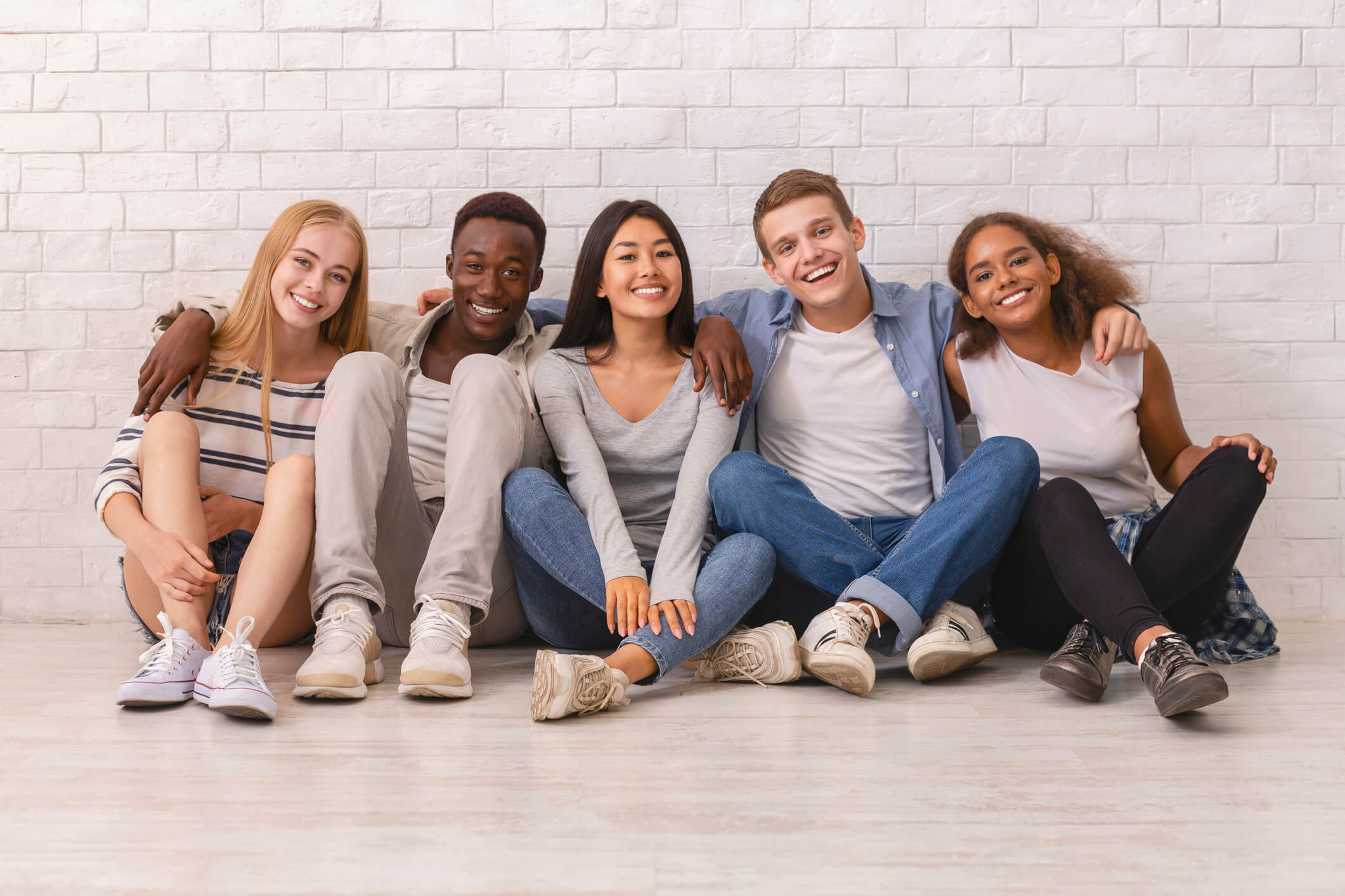 Diverse group of teens smiling and sitting together in front of a white brick wall