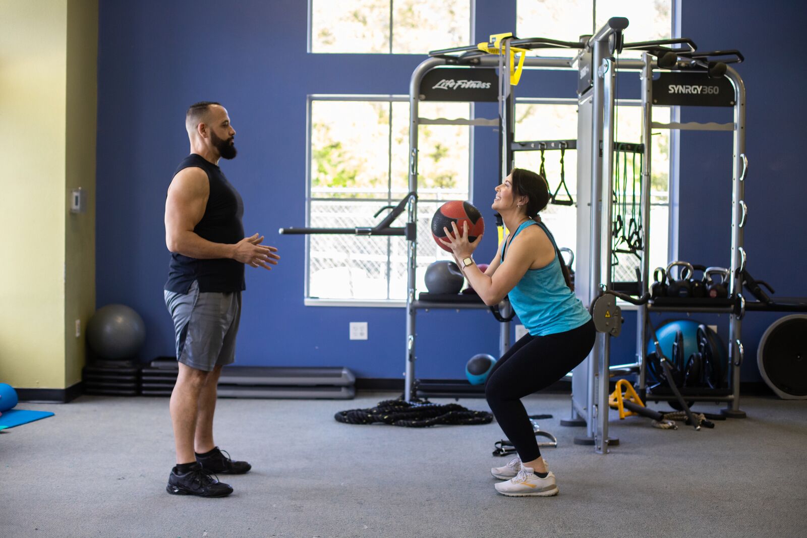 Image of man standing while woman is holding weighted medicine ball to throw to him as they stand in the gym's wellness floor