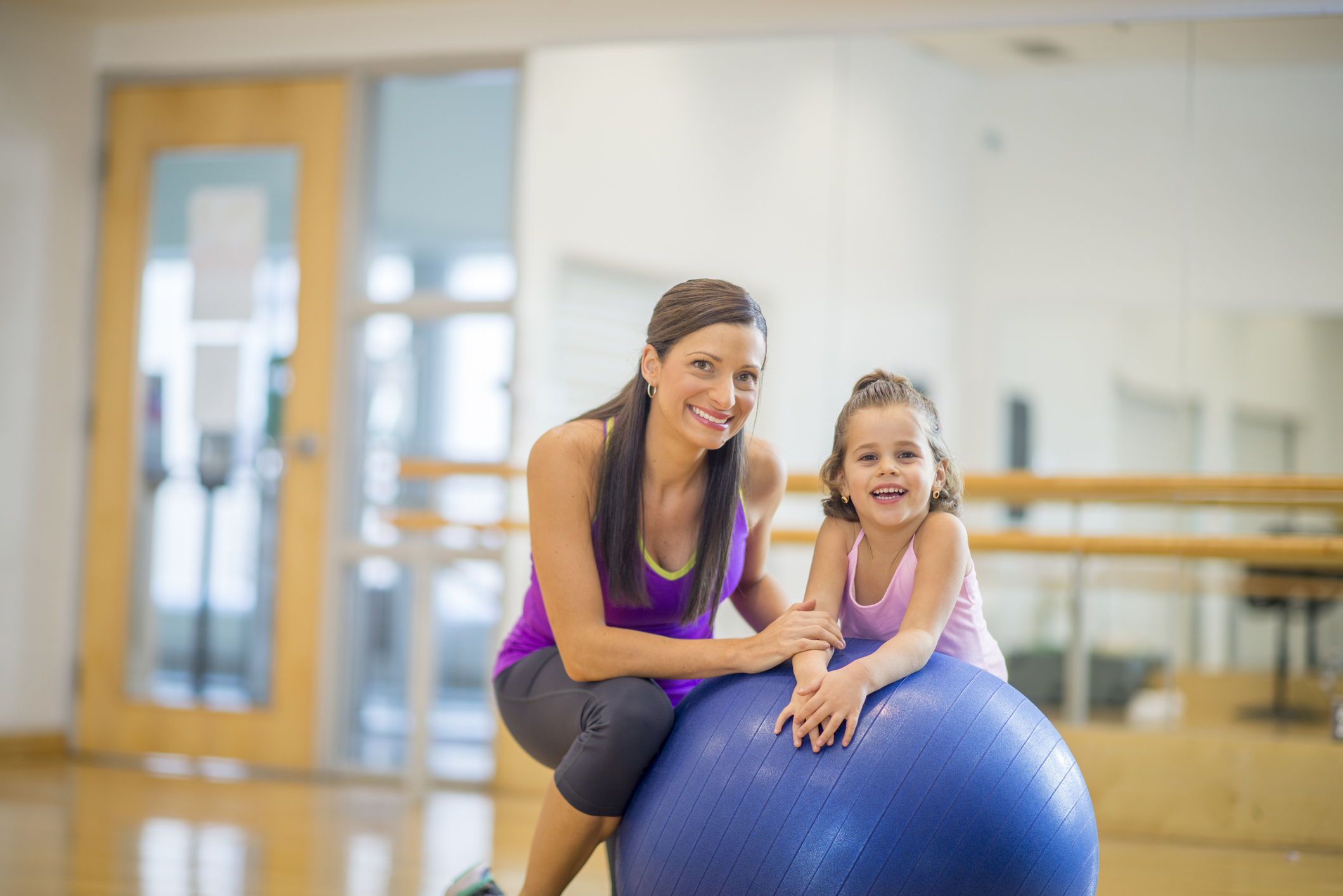 A mother and daughter are wearing athletic clothing and are happily together at the gym. They are smiling and looking at the camera.