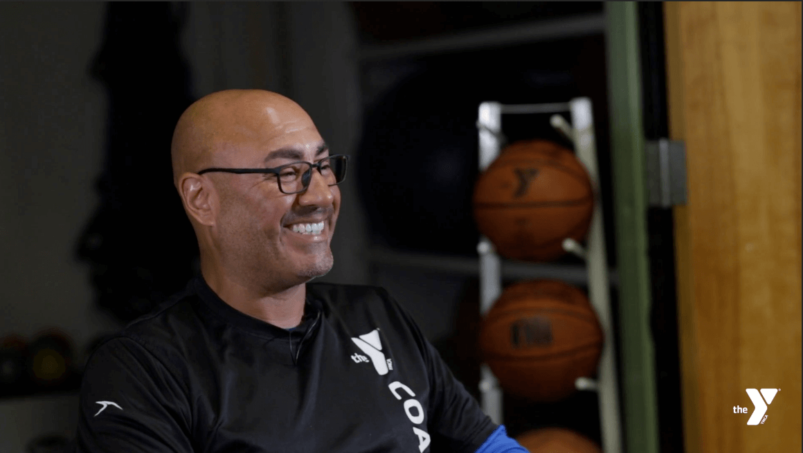 Image of Coach Ruiz sitting in front of basketballs wearing a black YMCA Coach shirt