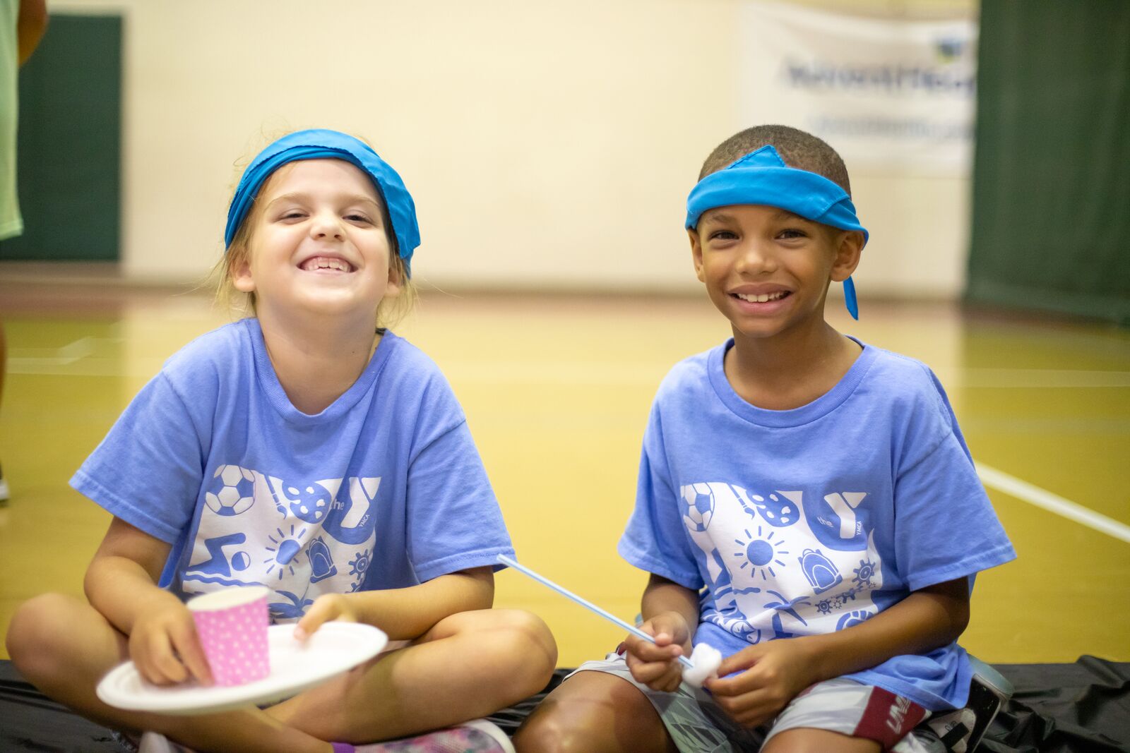 Two children wearing summer camp shirts while smiling in a gym and holding plates of lunch