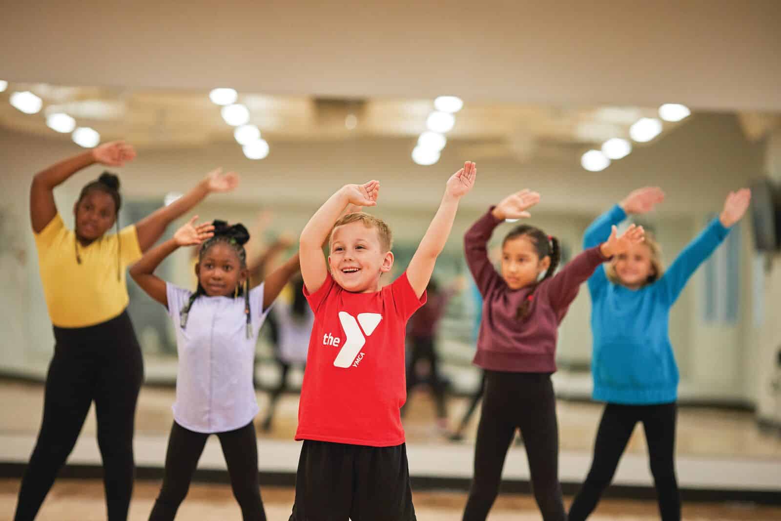 Kids smiling with their hands raised in the air dancing in a studio at the YMCA