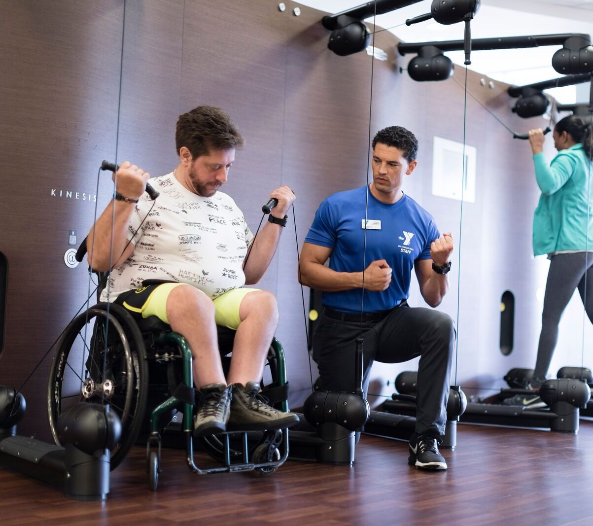 A member in a wheel chair using cable machines to workout with a trainer
